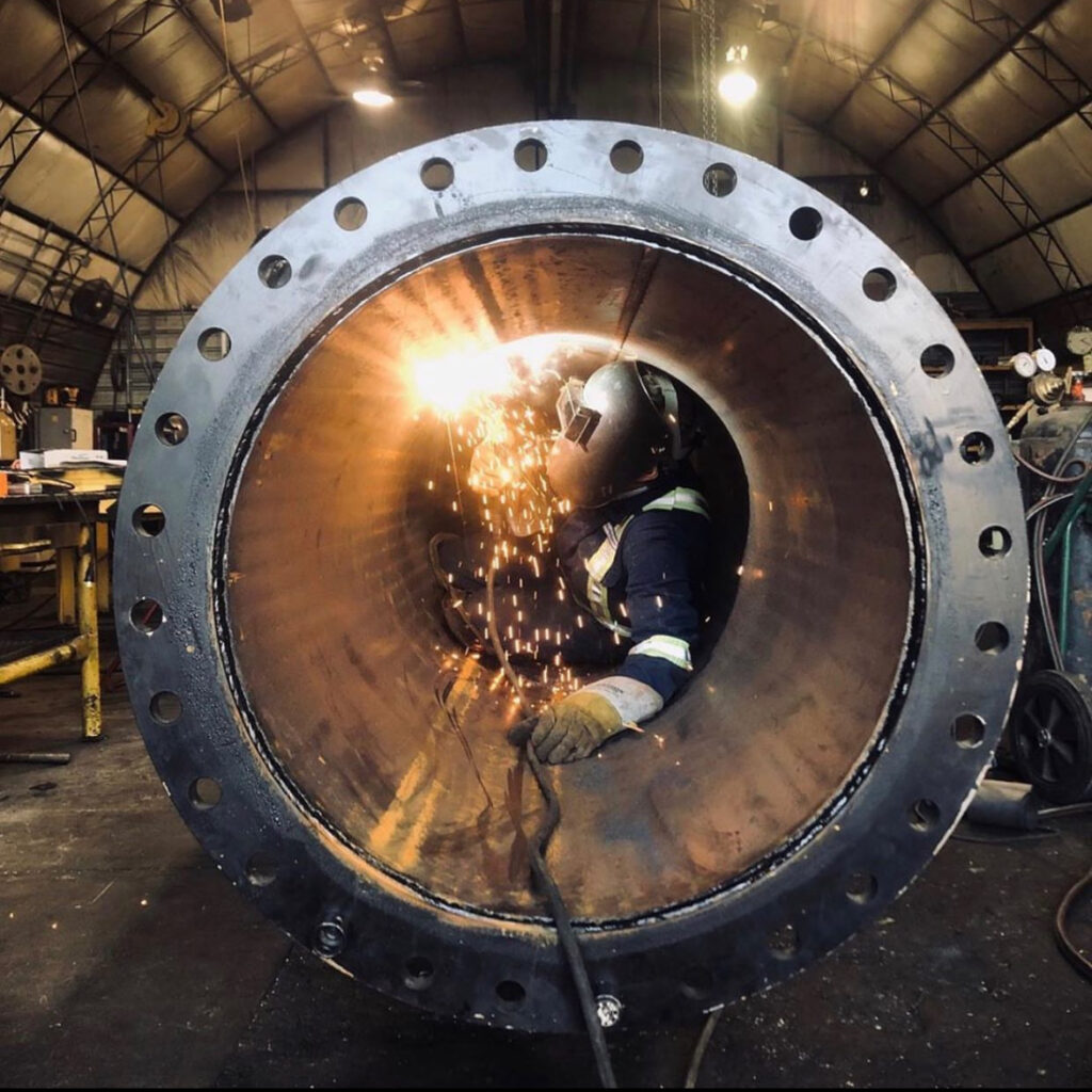 A welder in safety gear works inside a large cylindrical metal pipe, with sparks flying. The scene takes place in a spacious industrial workshop.