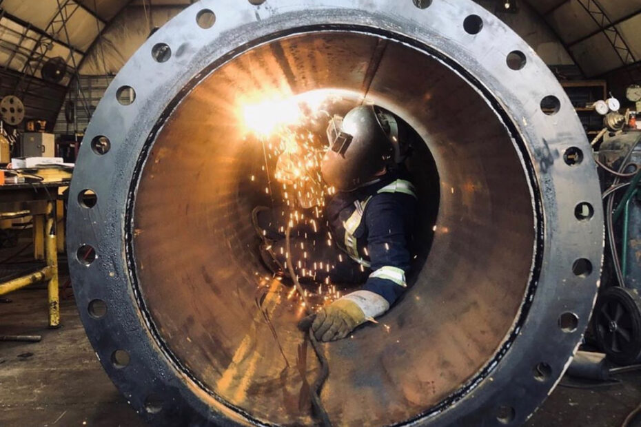 A welder in safety gear works inside a large cylindrical metal pipe, with sparks flying. The scene takes place in a spacious industrial workshop.